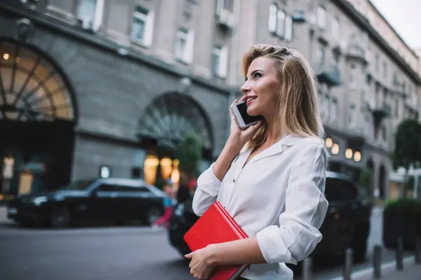 Side view of positive female student in trendy outfit holding notepad while walking along street and speaking on smartphone about project