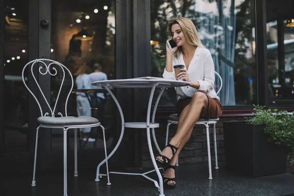Young positive female in casual clothes with toothy smile looking down while sitting at table on street with takeaway coffee and talking on smartphone