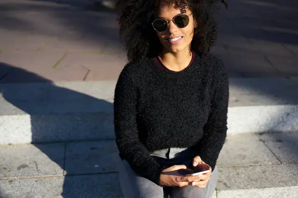 Crop young lady with curly hair in casual clothes and sunglasses resting on stairs of street and holding smartphone in sunny city
