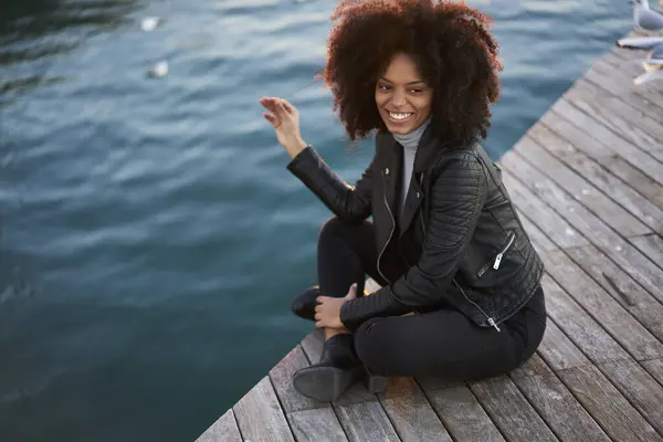 Full bosy of cheerful young African American female traveler with kinky hair in stylish outfit waving hand and looking away with toothy smile while having break on pier near lake