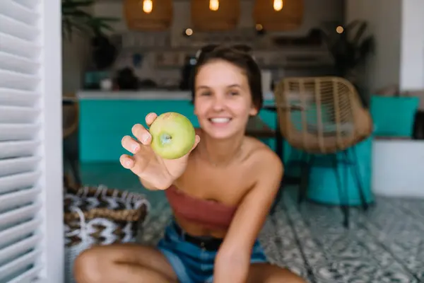 Soft focus of happy young fit female in casual outfit looking at camera and suggesting ripe green apple while sitting on floor in modern kitchen interior