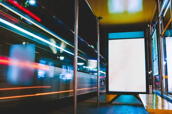 夜間にガラス壁と照らされた広告の旗が付いている空の駅の近くのぼやけた交通機関が付いている明るい通りの長い露出 ストックフォト