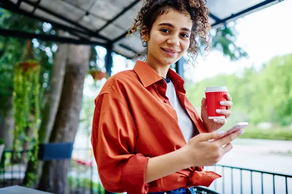休憩中に携帯電話でコーヒーとサーフィンインターネットのペーパーカップと立っている間 カールヘアを見ているクロップ笑顔のエスニック女性 ストック写真