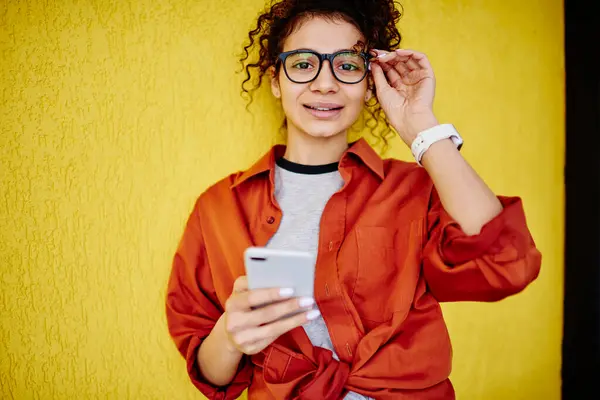 スマートフォンでイエローウォールサーフィンインターネットの近くに立っている間 カメラを見て 眼鏡のタッチフレームを微笑むエスニックな女性の作物 ストック写真