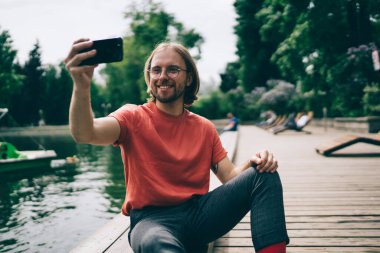 Keyifli sakallı, günlük kıyafetli, gülümseyen ve akıllı telefonu parktaki göl kenarında otururken selfie çekmek için kullanan adam.