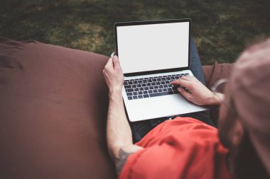 Şehrin parkında dizüstü bilgisayar kullanarak sosyal ağları kontrol eden bir erkeğin fasulye torbasının üzerinde dinlenmesi görüntüsü.