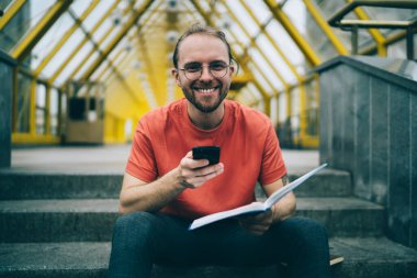 Gözlüklü mutlu adam kameraya bakıyor ve elinde akıllı telefon ve kitapla caddede gülümsüyor.