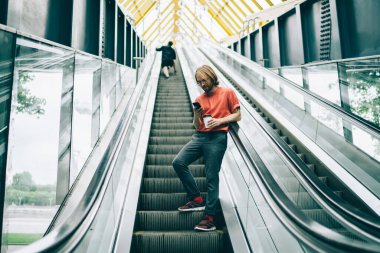 Sıradan bir erkek, gözlüklü, bir fincan kahve içiyor, akıllı telefonu kullanarak şehirde yürüyen merdivene yaslanmış arkadaşlarıyla mesajlaşıyor.