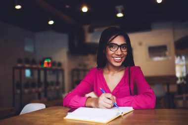 Gözlüklü ve dişlek gülümseyen mutlu Afrikalı Amerikalı kız öğrenci masada oturup not defterine notlar alırken gözlerini kaçırıyor.