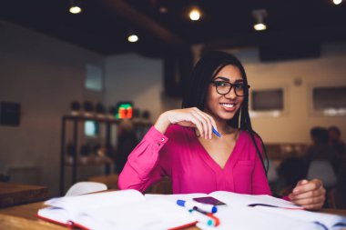 Gözlüklü ve dişlek gülümsemeli genç, mutlu Afro-Amerikan kız öğrenci masada oturmuş ödevlerini yaparken kameraya bakıyor.