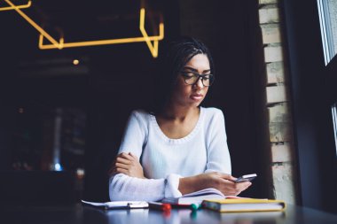 Gözlüklü, gözlüklü, elinde not defterleri ve ev ödevi yaparken cep telefonuyla masada oturan dalgın siyah kız öğrenci.