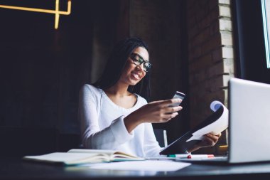 Güncel kıyafetlerle gezen, akıllı telefonlara bakan ve güncel iş yerinde belgelerle çalışırken gülümseyen Afro-Amerikan genç bir kadın.