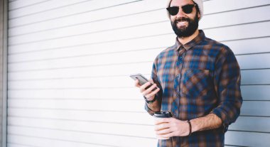 Gezintiler sırasında gülümseyen, cep telefonu teknolojisi ve kafein içeceği taşıyan neşeli hippi adamın yarım boy portresi. Cep telefonu ile mutlu bir Orta Doğulu erkek blogcu.