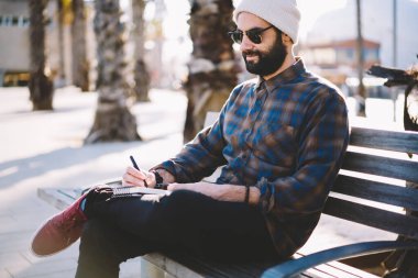 Güneş gözlüklü Türk hippi, seyahat tatillerinde solo ziyaret için yeni fikirler yazıyor. Ortadoğulu erkek öğrenci şehir bankında eğitim kitaplarını planlamak için kullanıyor.