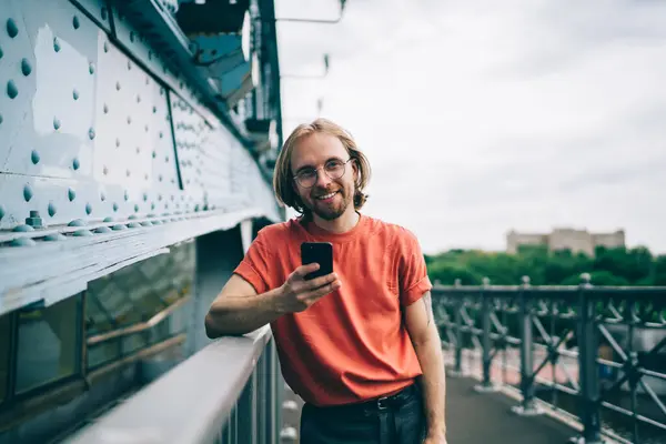 コンテンツ 陽気な赤ひげの若者 メガネで 長い髪 シャツ ブリッジに立っている携帯電話を見ている ロイヤリティフリーのストック画像