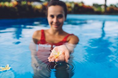 Kırmızı mayolu pozitif bir kadın boş havuzun berrak sularında duruyor ve palmiye üzerinde narin, sarımsı bir çiçek sergiliyor.