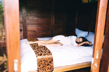 Beyaz gecelikli gülümseyen genç bir kadın güneş doğarken yatakta güneşleniyor ve tahta evde yastığa sarılıyor.