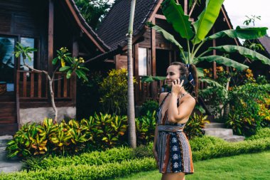 Yazın ahşap evin yakınındaki parlak yeşil bitkilerin yanından geçerken cep telefonuyla konuşan ve gülümseyen güzel elbiseli genç bir kadının yan görüntüsü.