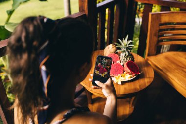 Yukarıdan gelen isimsiz bir kadın cep telefonunun ahşap yuvarlak masasında taze meyvelerin fotoğrafını çekiyor.