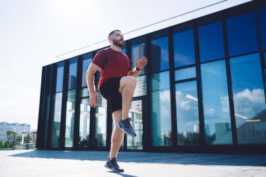 Güneşli bir sabahta spor ayakkabısı giymiş genç bir adamın modern ofis binasına karşı ısınması ve antrenmanın keyfini çıkarması ve başka yere bakması.