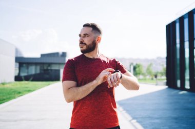 Kırmızı tişörtlü, kendinden emin bir erkek şehir sokağının bulanık arka planında dikilirken bileğinde modern akıllı saati kullanırken başka tarafa bakıyor.