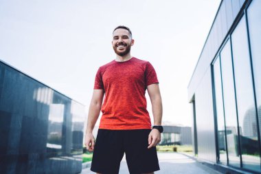 Spor giyim sektöründe düşük açılı kaslı bir erkek koşucu şehrin caddesinde durup kameraya bakarken gülümsüyor.