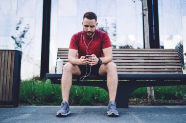 Spor giyim sektöründe ciddi sakallı bir adam müzik dinlerken ve şehir parkında yedek kulübesinde vakit geçirirken cep telefonunu karıştırıyor.