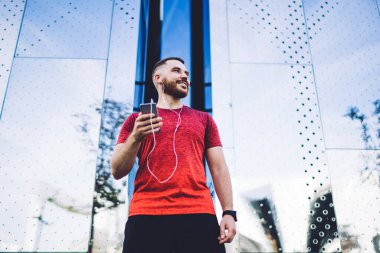 Kırmızı gömlekli, kulaklıklı, cep telefonlu mutlu bir genç modern binanın metal duvarının yanında dikilirken...