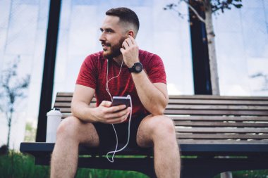 Şık saç kesimi olan neşeli sakallı erkek ahşap bankta dinlenirken akıllı telefon tutuyor ve eğitimden sonra kulaklıkla müzik dinliyor.