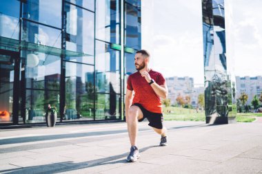 Spor ayakkabılı güçlü sporcu vücudu kaldırımlı caddede bacaklarını esneterek antrenman yapıyor ve başka tarafa bakıyor.