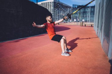 Uzun boylu, etkin erkek sporcuların güneşli yaz günlerinde spor sahasında dikilirken süspansiyon antrenmanı yapmasının yan görüntüsü.