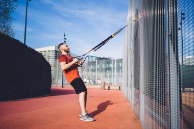 Çitin yanında durup güneşli yaz günlerinde okul bahçesinde süspansiyon eğitimi alan aktif giyimli güçlü bir erkek sporcunun yan görüntüsü