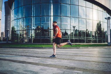 Gündüz vakti modern cam şehir binasının yanındaki kaldırımda koşan spor kıyafetleriyle koşan tüm erkek koşucunun yan görüntüsü.