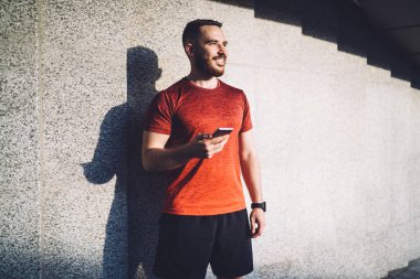 Neşeli, sakallı, spor kıyafetleri giyen genç bir erkek atlet taş duvarın yanında durup gülümseyerek gözlerini kaçırıyor.