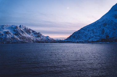 Norveç fiyortlarının kayalık oluşumlarının resimli manzarası alacakaranlıkta akşam vakti dalgalanan denizin yanındaki karı kaplıyordu.