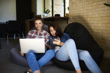 Gayri resmi giyinmiş genç kadınlar, modern çalışma alanında birlikte plan yaparken fasulye çuvallarının üzerinde oturup dizüstü bilgisayar ve tablet kullanıyorlar.