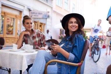 Motosikletli bir kadın sokakta bisiklet sürerken arkadaşlarıyla açık kafede kahve içerken moda şapka sörfü tabletinde pozitif bir kadın.