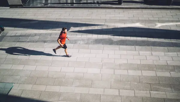 From above of full body young male athlete running on pavement in city and looking away while living healthy lifestyle and warming up in morning