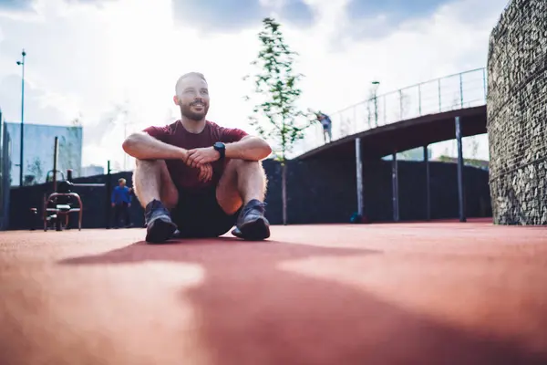 スニーカーで笑顔のひげ付きスポーツマンが地面に座って 晴れた朝の激しいトレーニングの後に休んでいる間 遠ざかる全身 ロイヤリティフリーのストック画像