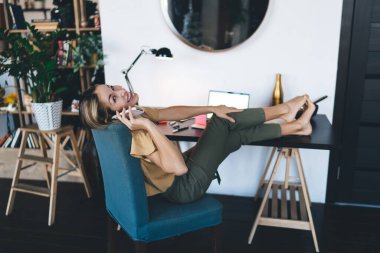 Avrupalı bir kız evde cep telefonuyla konuşuyor. Modern kadın kavramı. Uzaktan veya e-öğrenme fikri. Genç, neşeli bir kadın, açık bir dizüstü bilgisayarla koltukta çıplak ayakla oturuyor. Stüdyo daire içi