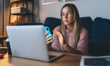 Gözlüklü Avrupalı kız evde cep telefonu kullanıyor. Ev hayatı kavramı. Ev istirahatı ve boş zaman fikri. Masada dizüstü bilgisayarı açık oturan genç bir kadın. Stüdyo dairenin içi.