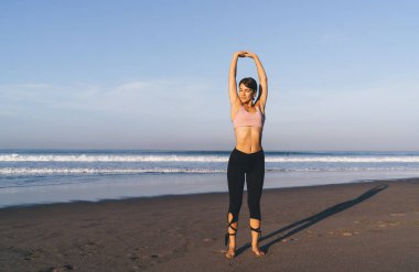 Sahil şeridinde güneş ışığı altında esneyen eşofmanlı genç bir kadın sportif ısınma için zaman harcıyor, beyaz bir kadın spor yaparken vücut esnekliği için ellerini kaldırıyor.