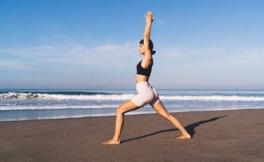 Aktif giyinen beyaz bir kadın sahil şeridinde egzersiz yapıyor vücut şeklini mükemmel tutuyor, esnek bir kadın okyanus kenarında sabah yogası yapıyor meditasyondan keyif alıyor.