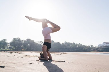 Atletik vücudu olan güçlü bir kadın Asya 'da ellerinin ve kafasının üzerinde duruyor kaslarını esnetiyor, sabahları deniz kenarında yoga meditasyonu yapıyor sağlıklı yaşam tarzını fiziksel sağlık için koruyor.