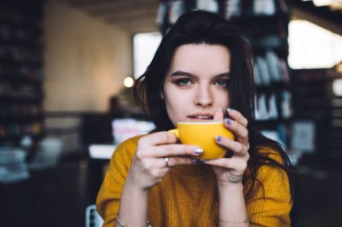 Modern iş yerinde kahve içerken sıcak içkinin tadını çıkaran ve kameraya bakan modaya uygun kıyafetleri olan şehvetli genç bir kadın.