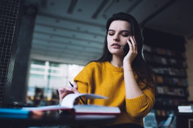 Ciddi bir genç asistanın cep telefonuyla konuşan ve kitap masasında otururken not alan sıradan bir Jumper 'dan.