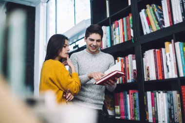 Sıcak süveterli genç adam kütüphanede ödevlerini yaparken konsantre olmuş sınıf arkadaşının kitap seçmesine yardım ediyor.