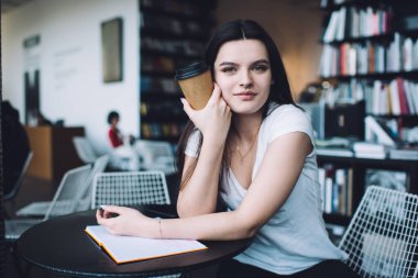 Günlük kıyafetli genç bayan kafasının yanında kahve fincanı ile masada oturuyor ve modern çalışma alanı projesinde çalışırken kameraya bakıyor.