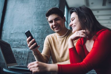 Samimi çalışma alanında netbook 'la masada otururken kırmızı kazaklı genç kız arkadaşıyla cep telefonu sörfü yapan pozitif adam.