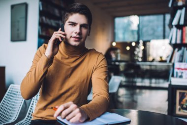 Düşünceli genç erkek, yaratıcı bir yerde açık bir defterle masada otururken cep telefonuyla konuşuyor ve kameraya bakıyor.